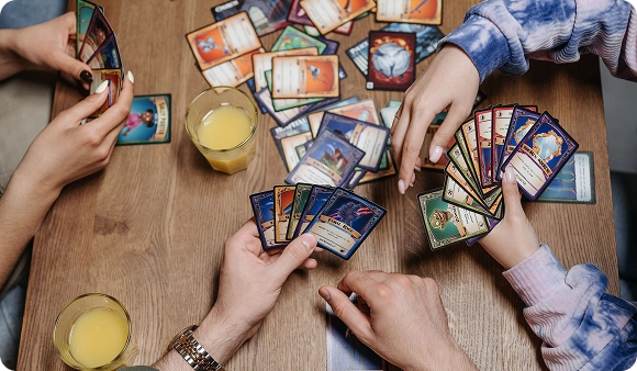 Um grupo de amigos, sentados à volta de uma mesa de madeira, segura leques de cartas enquanto jogam um jogo de cartas de mesa